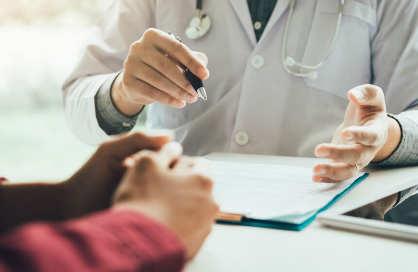 Healthcare provider in a white lab coat with a stethoscope gestures while speaking with a patient across a desk, with paperwork on a clipboard between them.