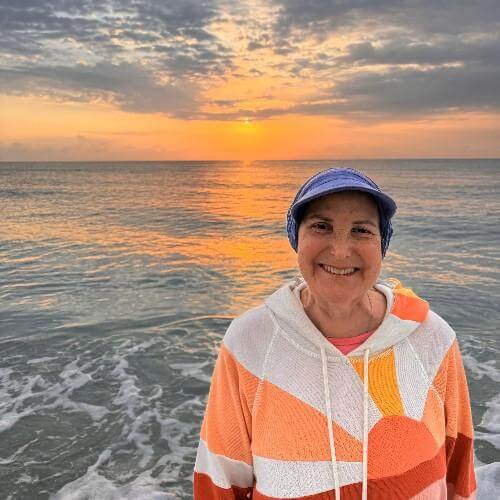 Pam Worth wearing a colorful orange hoodie and a jaunty blue hat in front of the ocean.