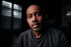 A portrait of Mike Cullen Johnson set against a dark background. He's wearing a grey t-shirt and looking at the camera.