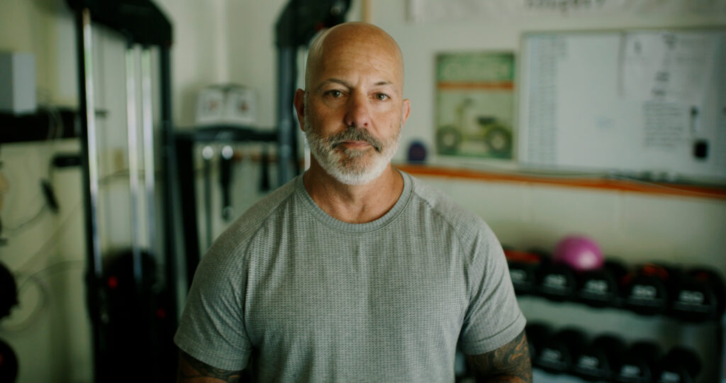 A portrait of Eric Gelber in his gym wearing a grey t-shirt and looking at the camera